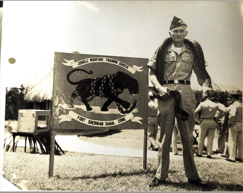 A smiling uniformed male soldier stands in front of a sign for the Jungle Warfare Training Center at Fort Sherman, Panama Canal Zone, with a python draped around his neck.